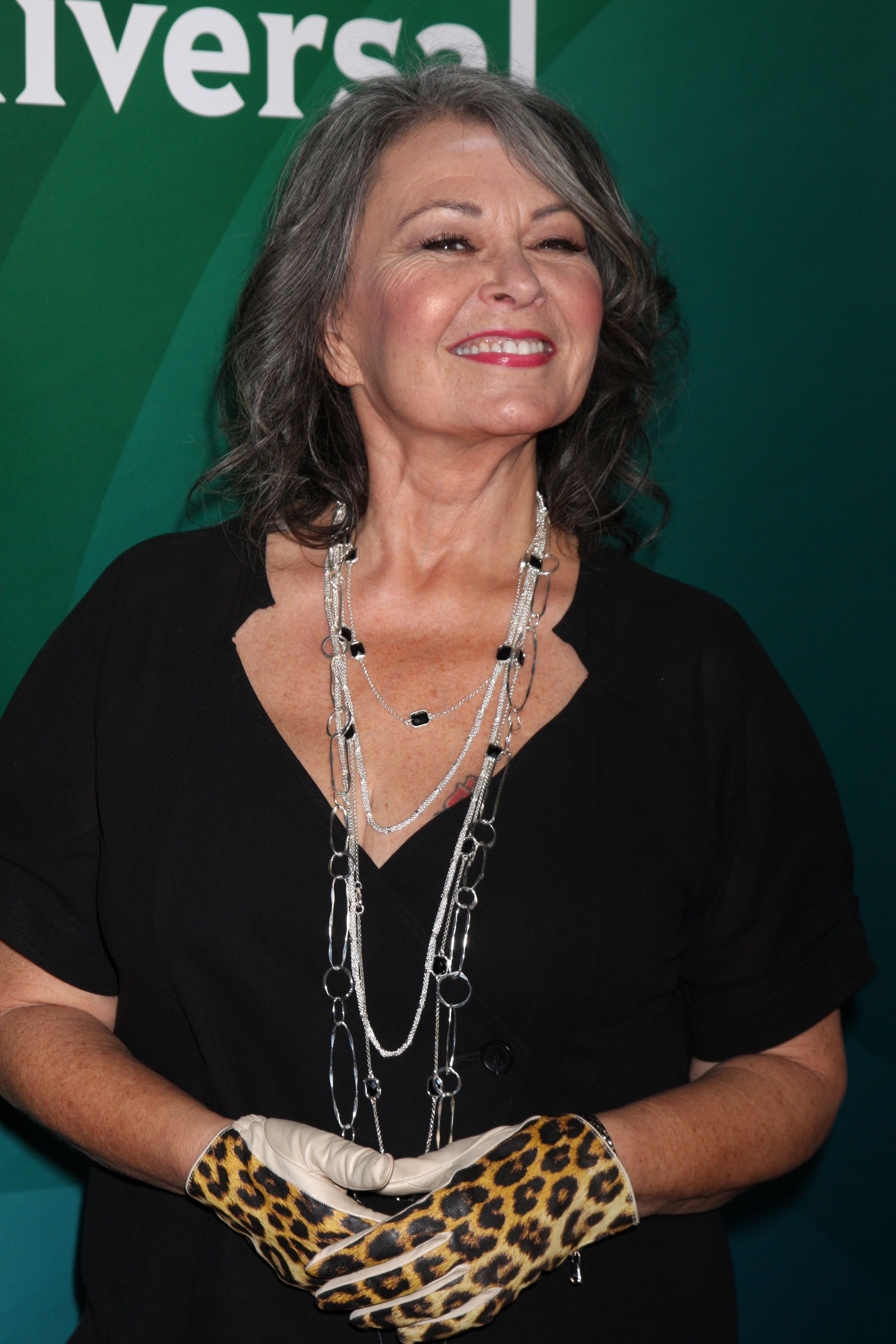 Roseanne Barr smiles for the camera as she wears a black top, statement chain necklace, and leopard-print gloves at an event.