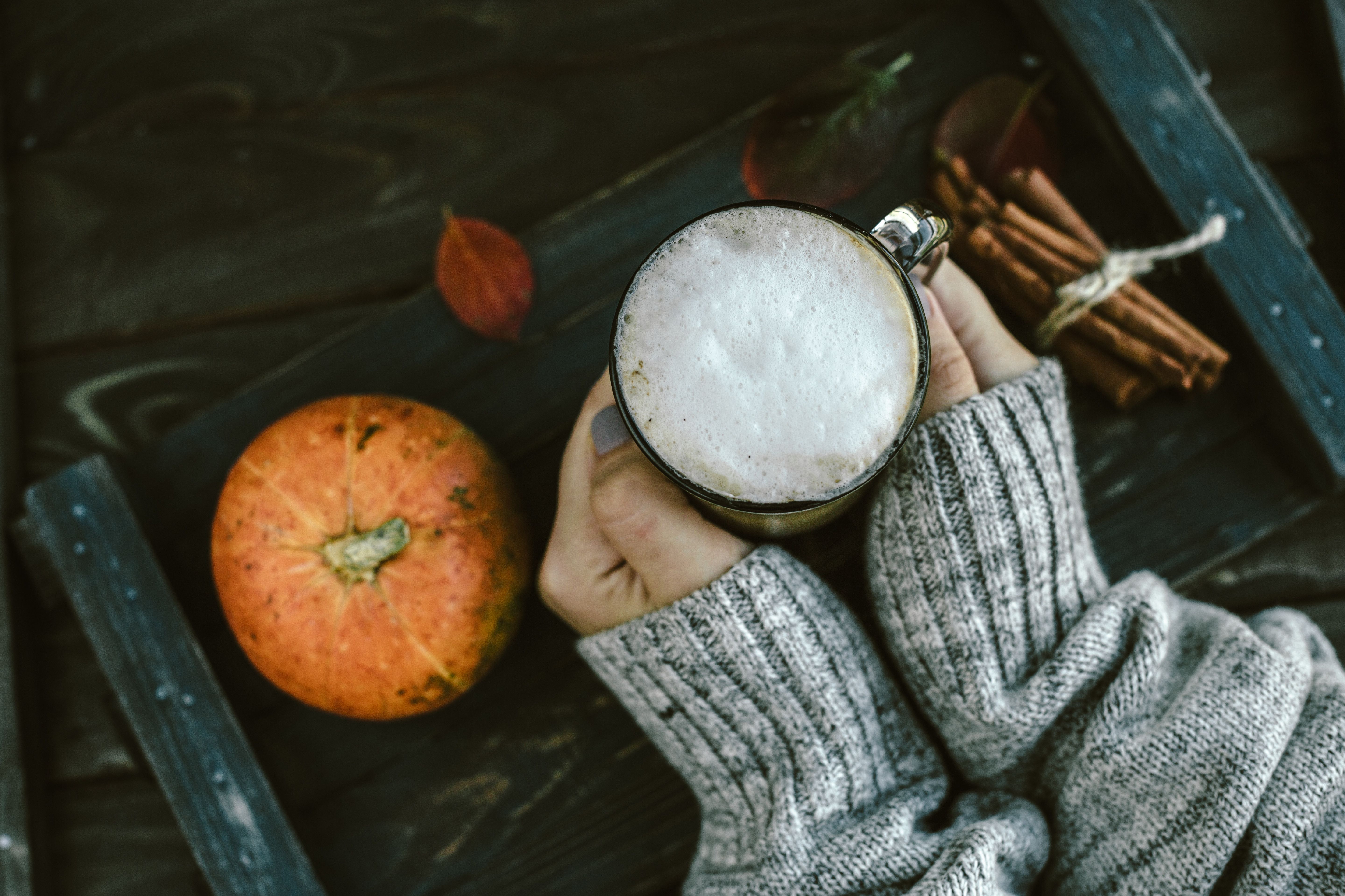 Woman wearing gray sweater holds palms around a latte on a tray with pumpkin, cinnamon sticks and autumn leaves. 