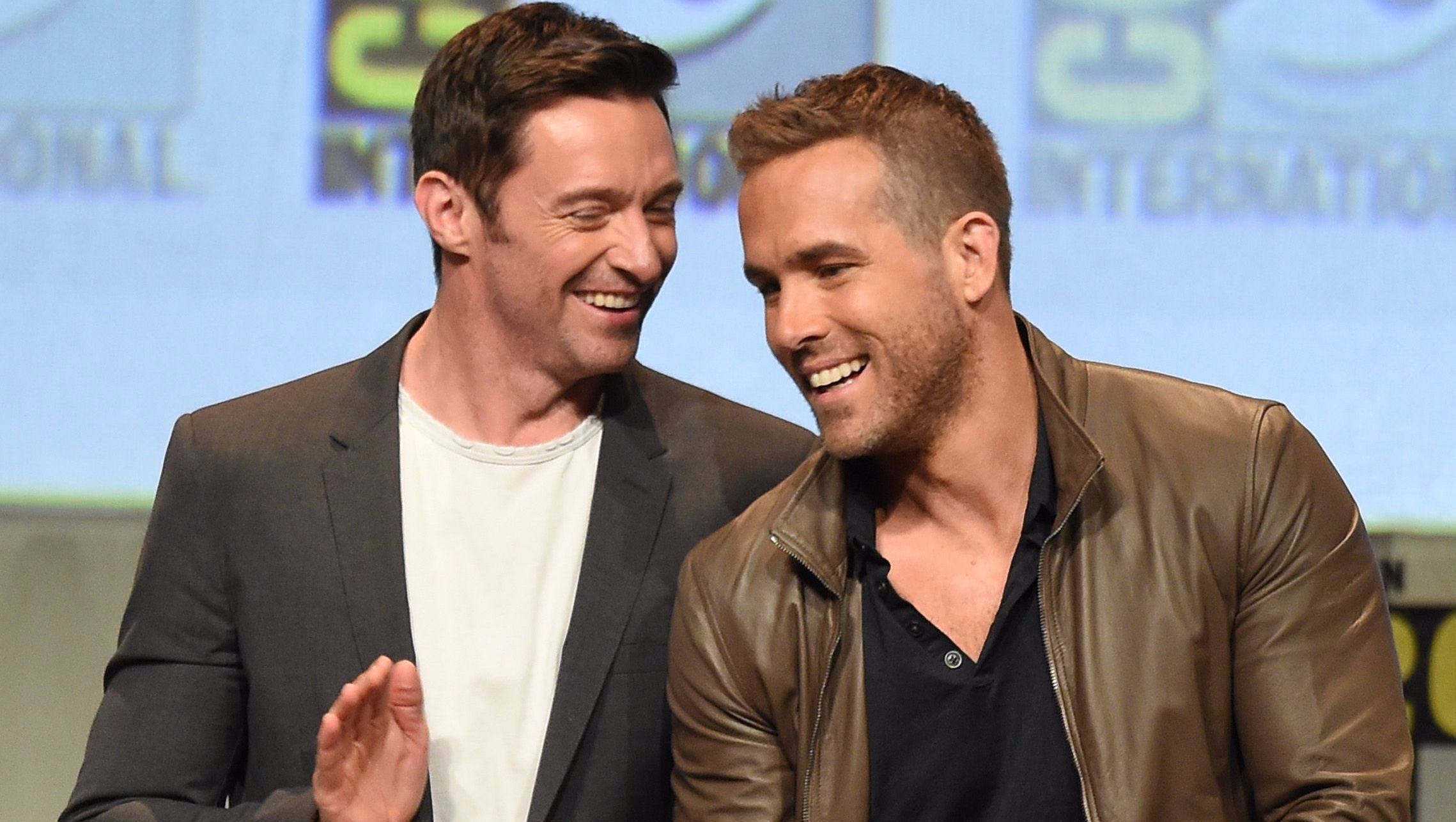 Hugh Jackman laughs with Ryan Reynolds in a brown jacket.