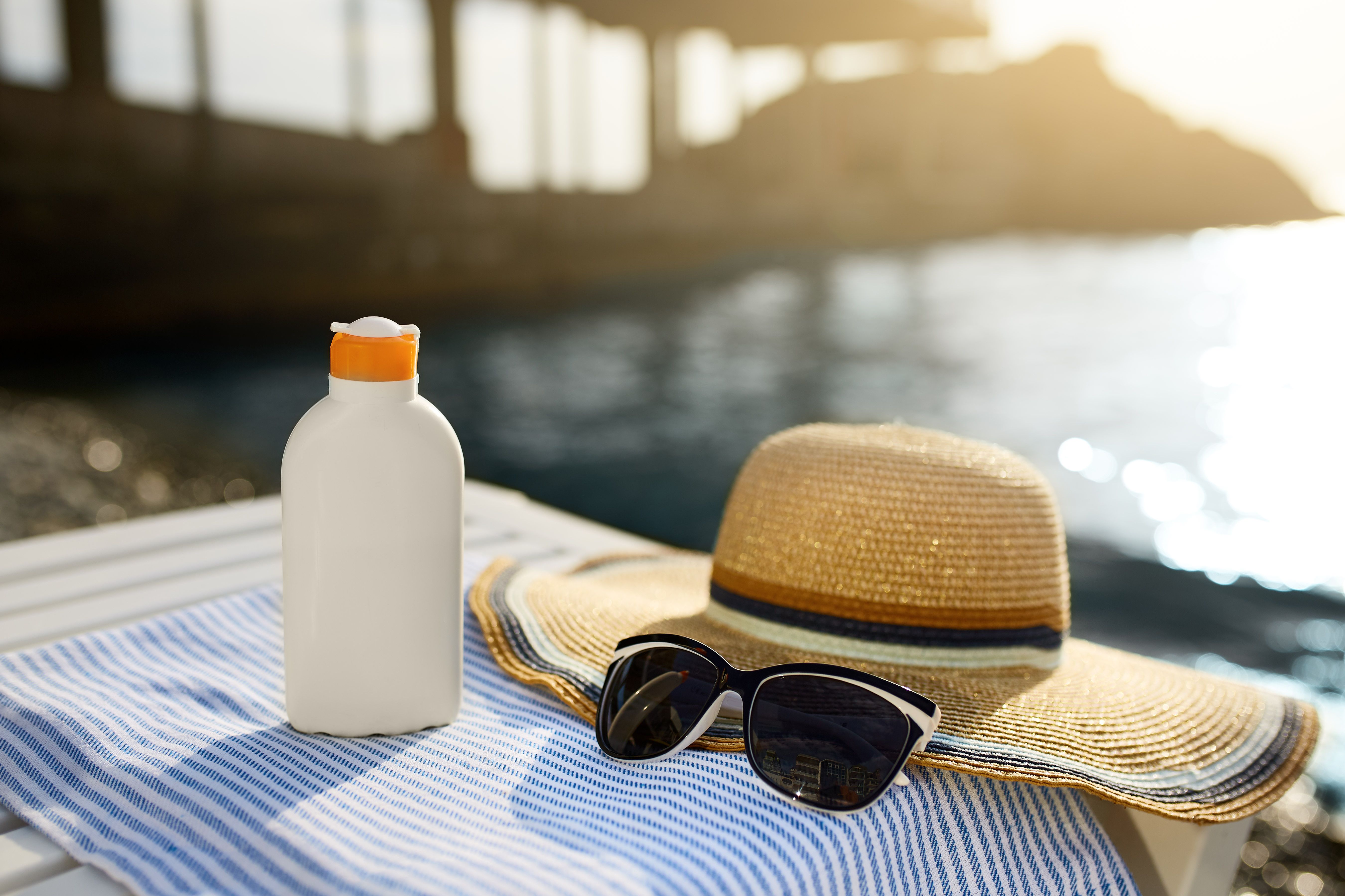 Sunscreen bottle on striped towel next to sunglasses and straw hat.