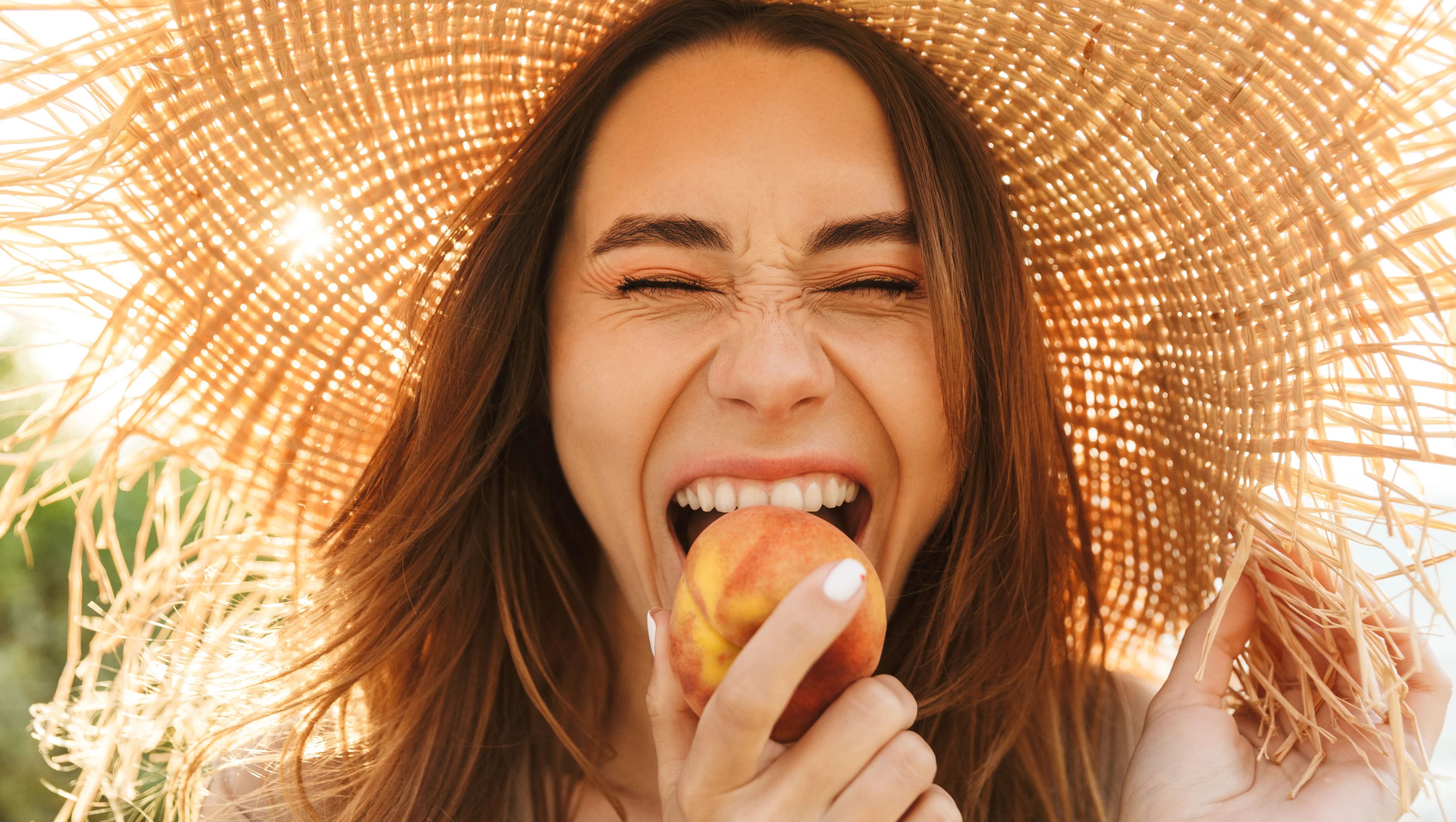 Young woman wearing straw hat bites into a peach. 