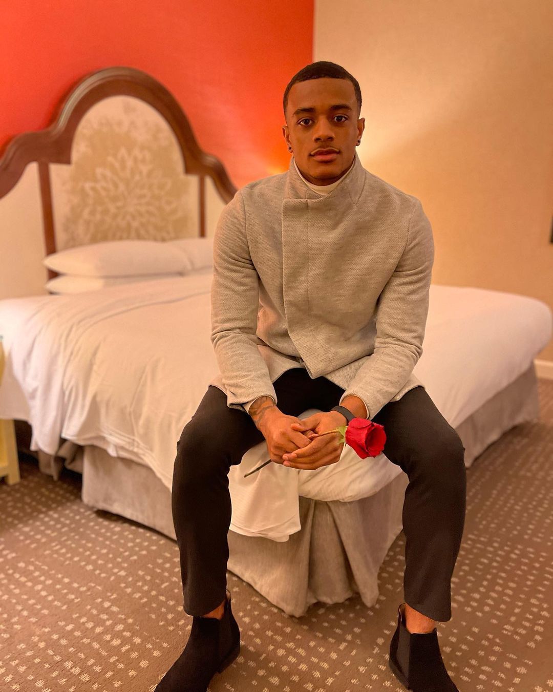 Patrick Surtain II casually sitting on a bed holding a long stem red rose.