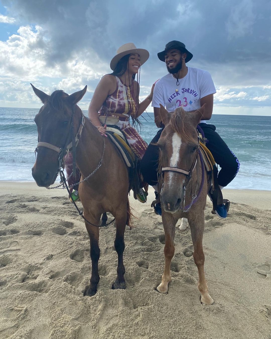 Jordyn Woods and Karl Anthony-Towns sit horseback side by side at the beach. 