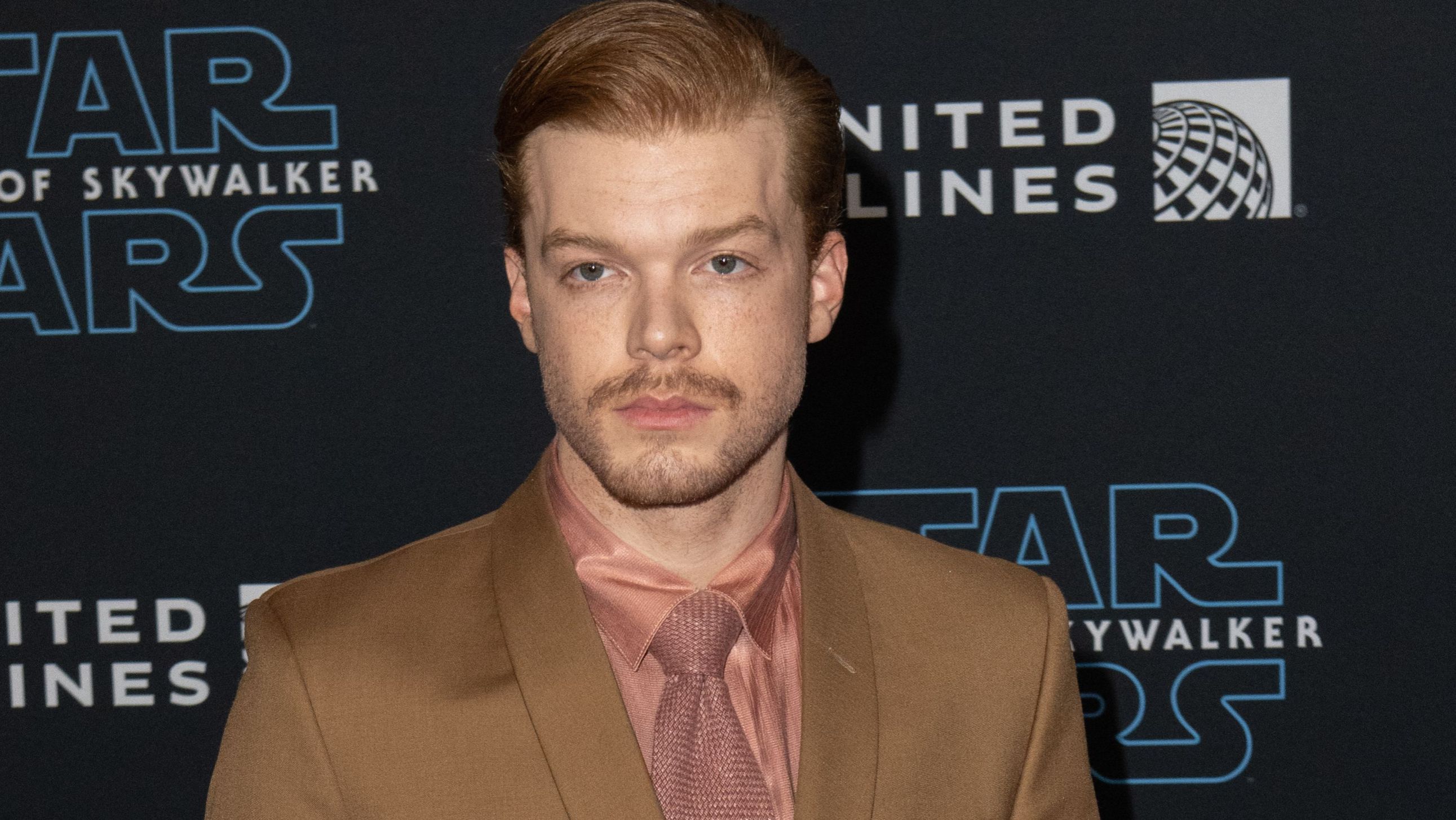Cameron Monaghan wears a tan suit and orange shirt.
