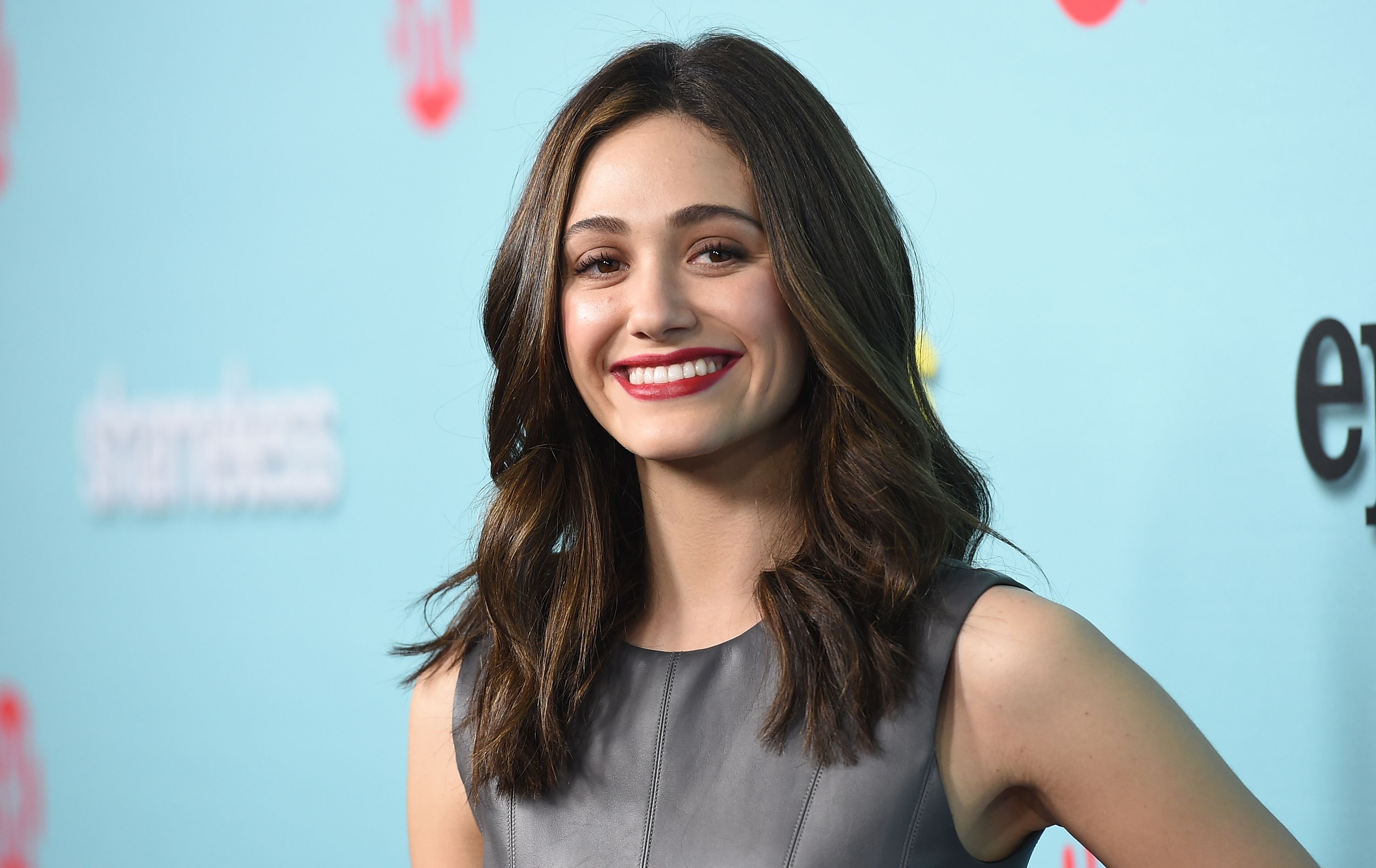 Emmy Rossum smiles with red lipstick and a black dress.