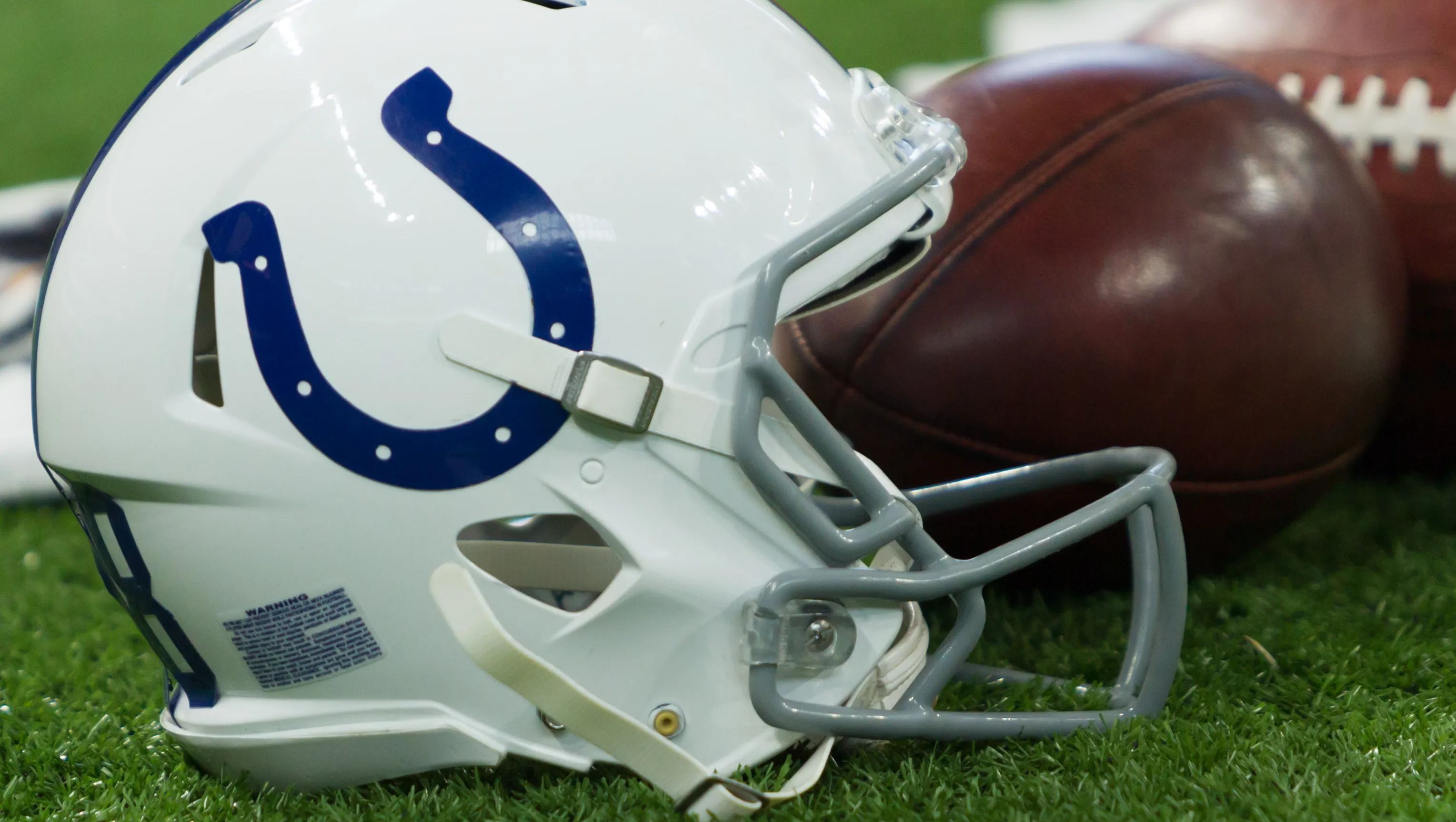 An Indianapolis Colts helmet next to two footballs on the field. 