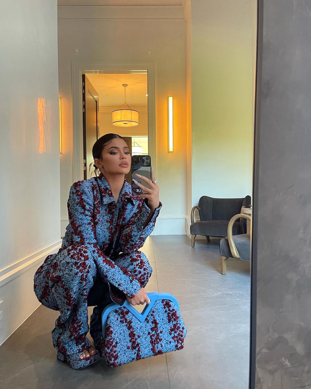 Kylie Jenner takes a selfie in patterned blue jumpsuit, matching bag, and heels. 