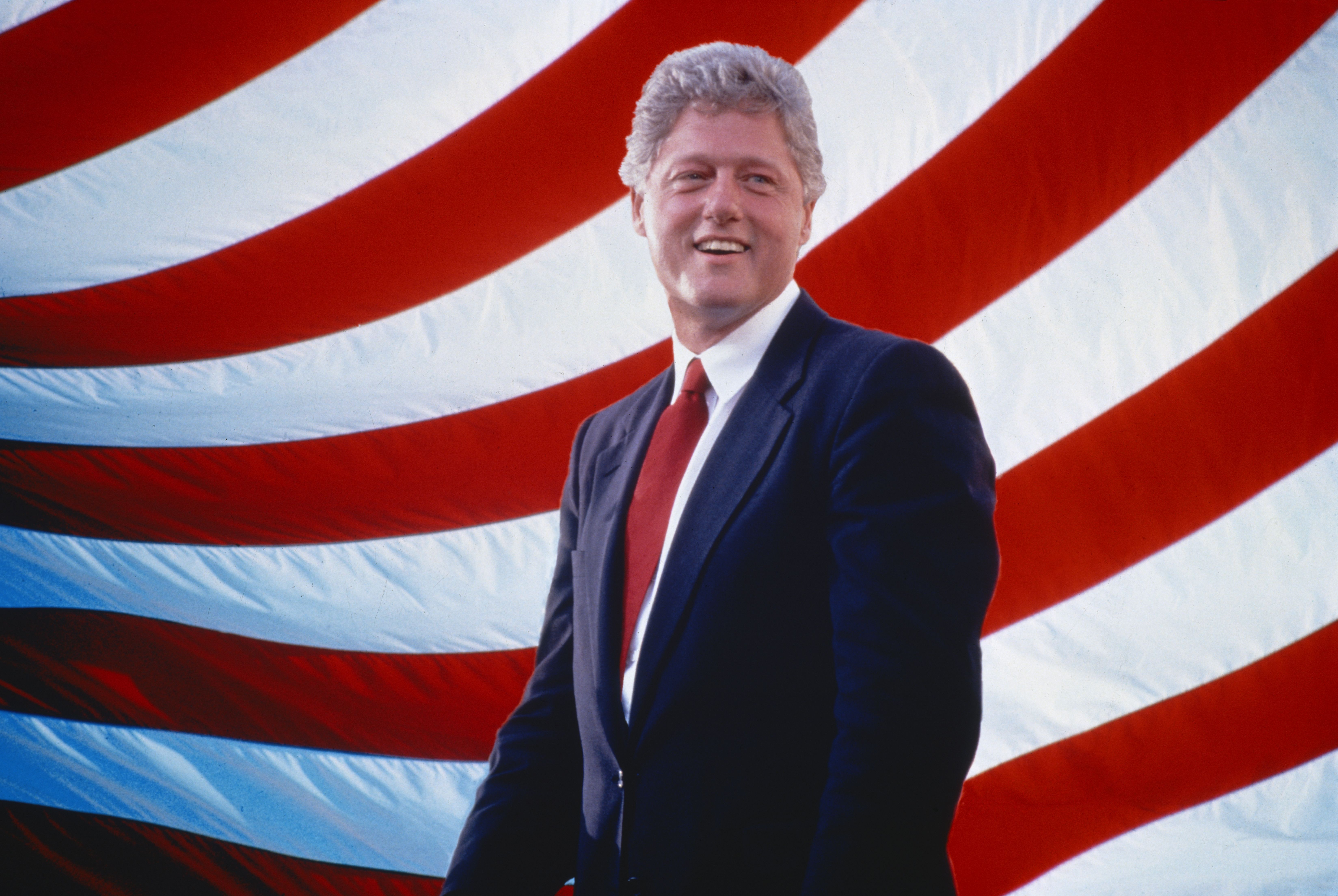 Bill Clinton stands in a blue suit in front of the American flag.